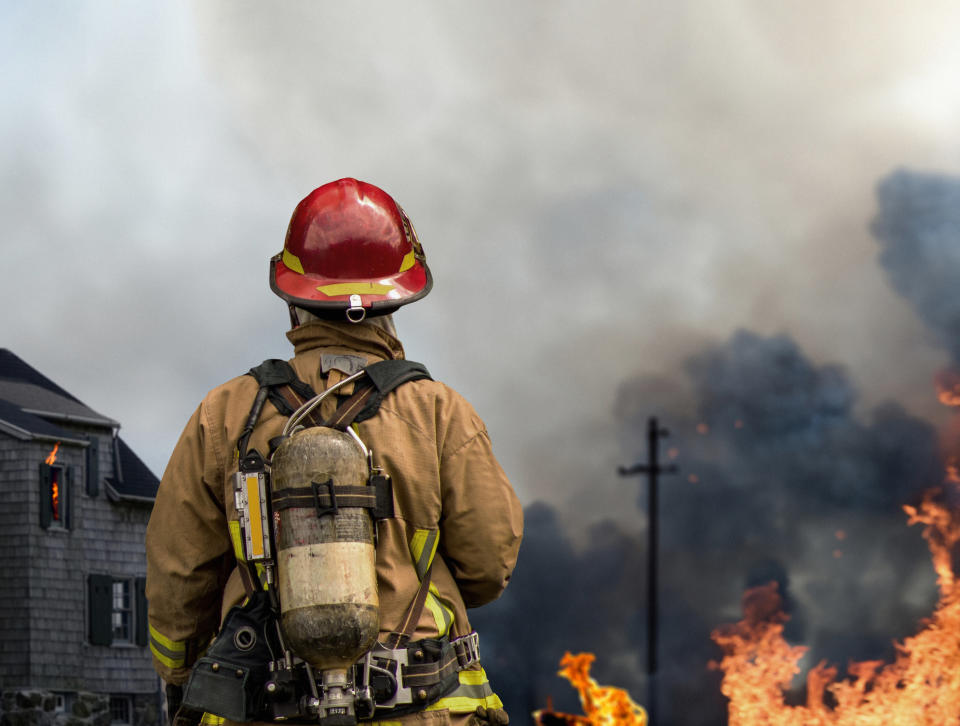 Firefighter in full gear facing a building engulfed in flames and smoke