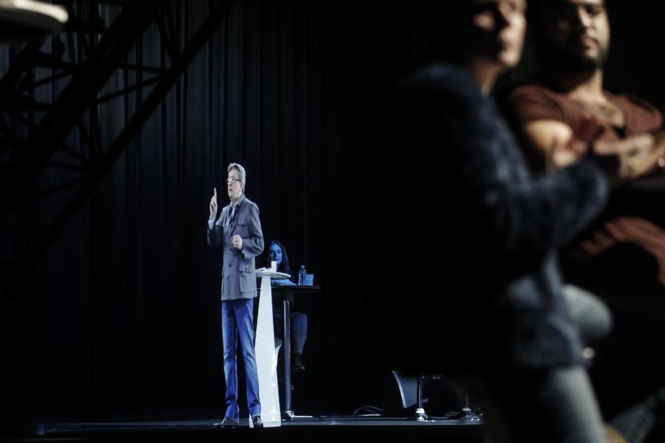 People sit at a table on stage and look towards the hologram of hard-left French presidential candidate Jean-Luc Melenchon, as he speaks to supporters who are gathered in Saint-Denis, near Paris, Sunday, Feb. 5, 2017. As Melenchon holds a rally in Lyon Sunday, a hologram of him is being projected by satellite to crowds in Paris. (AP Photo/Kamil Zihnioglu)