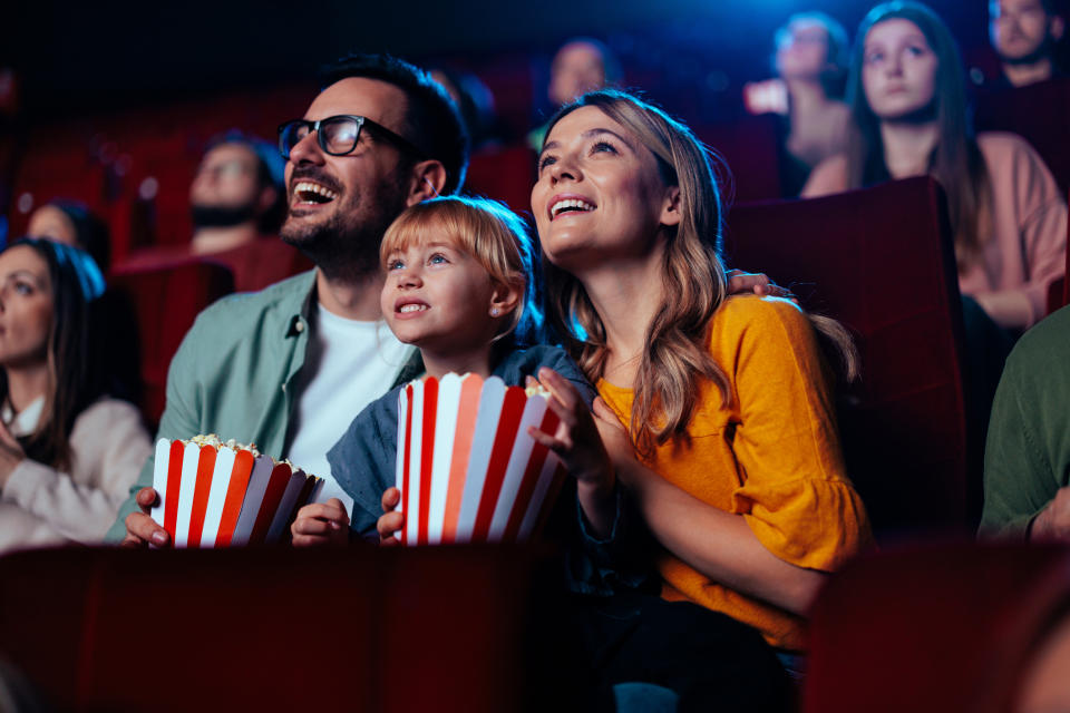 A family of three, with parents and a young child, happily watching a movie in a theater, holding buckets of popcorn. Other audience members are in the background