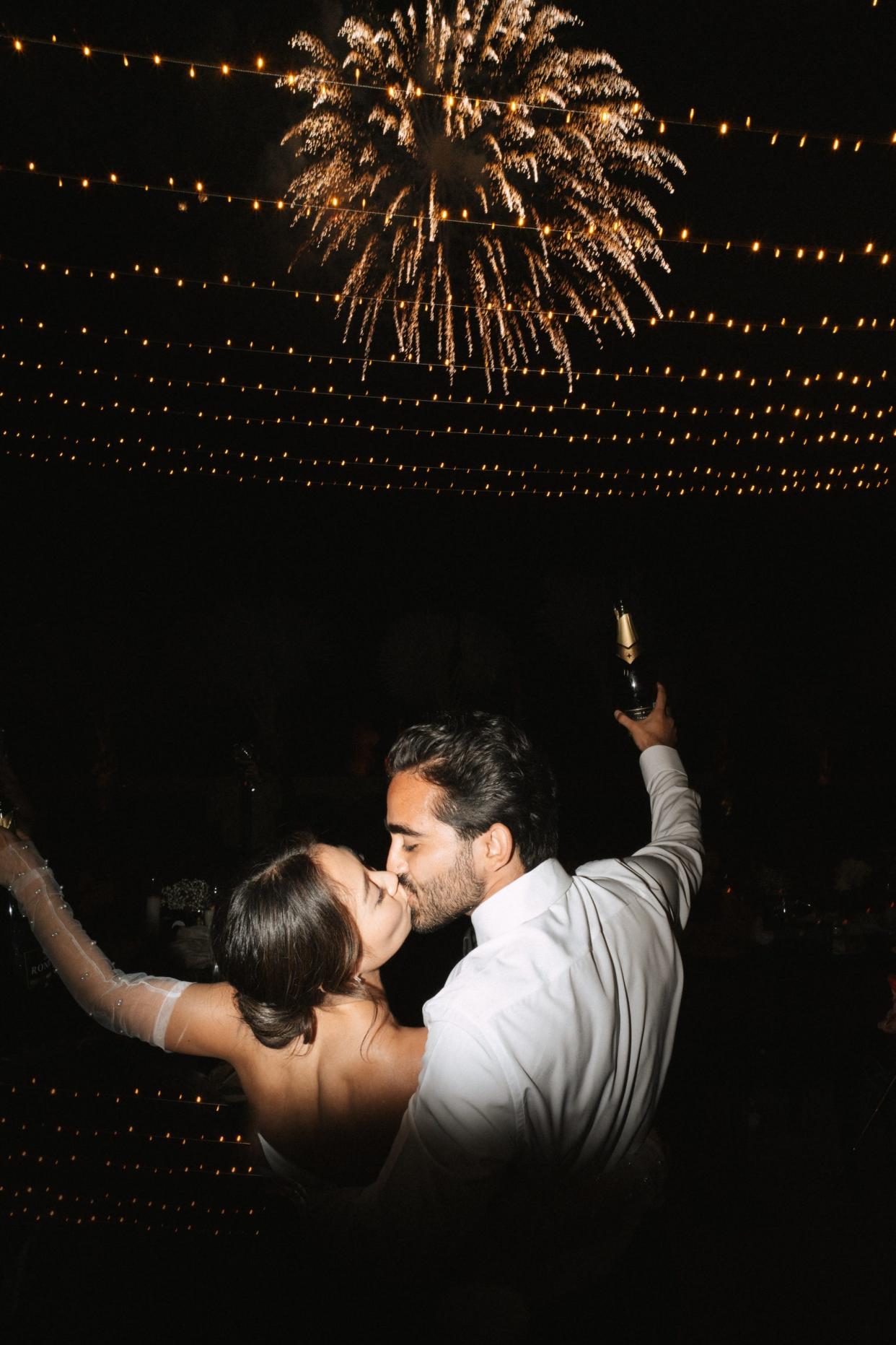 A couple kisses in front of fireworks and raise glasses of champagne. 