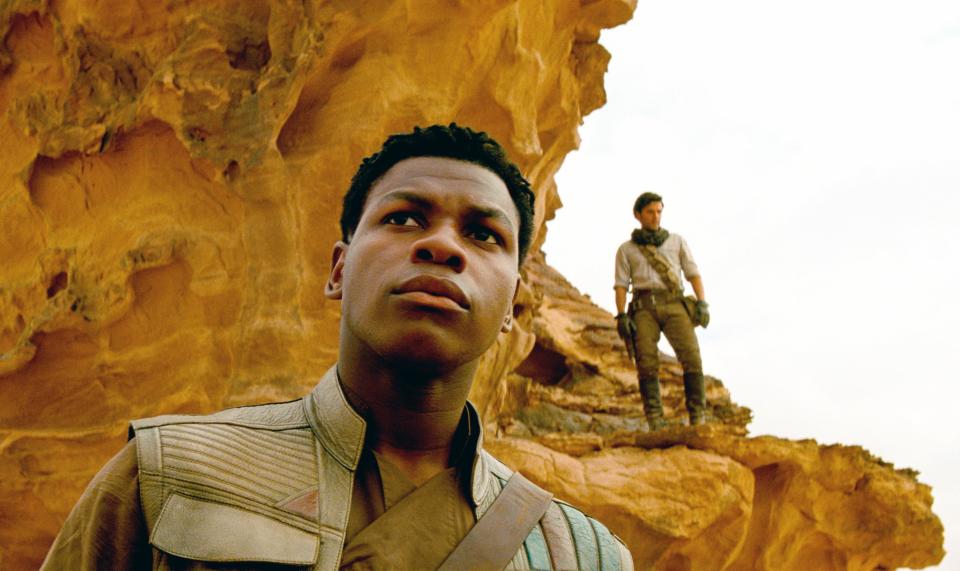 John Boyega and Oscar Isaac, in Star Wars costumes, stand on a rocky terrain set from a desert planet. Boyega looks ahead, Isaac stands in the background