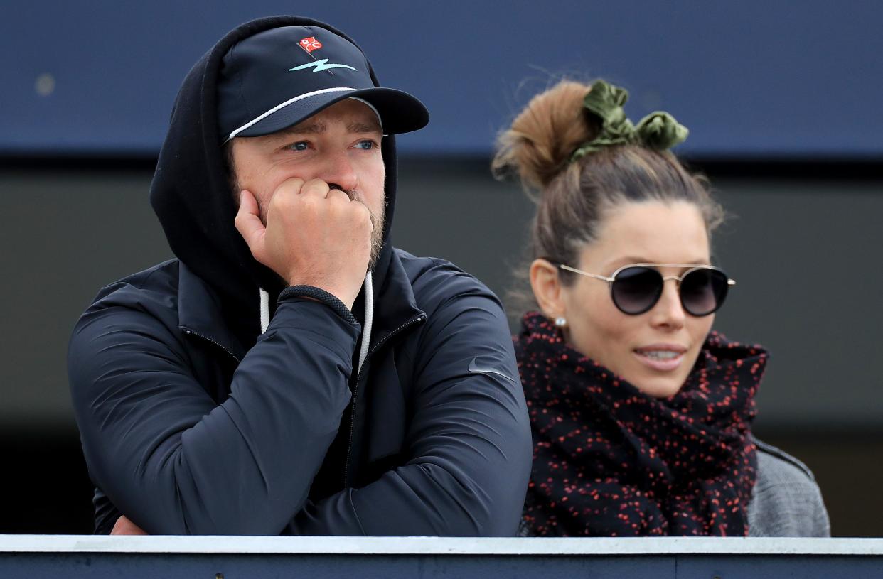 Justin Timberlake is on the left wearing a hoodie and resting his head on his hand. Jessica Biel is on the right wearing sunglasses. They are watching a game.