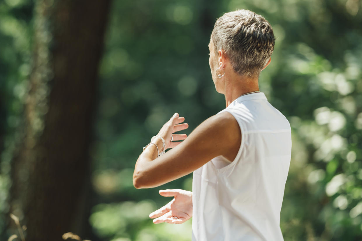 A short-haired woman in white sleeveless top, with back to camera, in white practices tai chi.