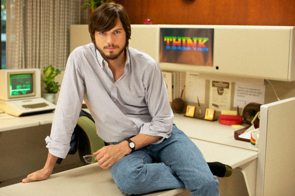 Ashton Kutcher as Steve Jobs, sitting and posing on a desk