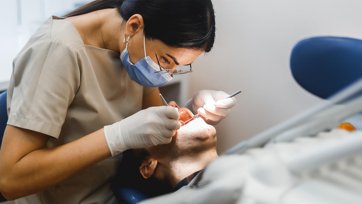  Dentist wearing a blue surgical mask and gloves leans over a patient's head to inspect his teeth with a mirror. 