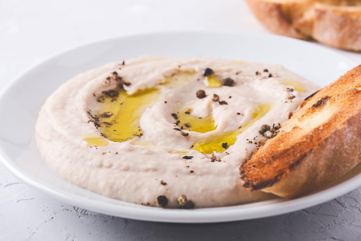 English muffin with a white bowl of hummus with a blurred background of English muffins