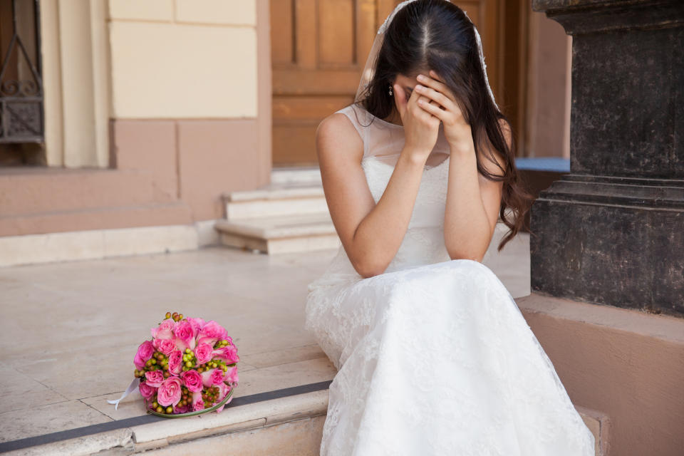 Bride in a white lace gown and veil sits on stone steps, covering her face with her hands. A bouquet of pink roses lies beside her