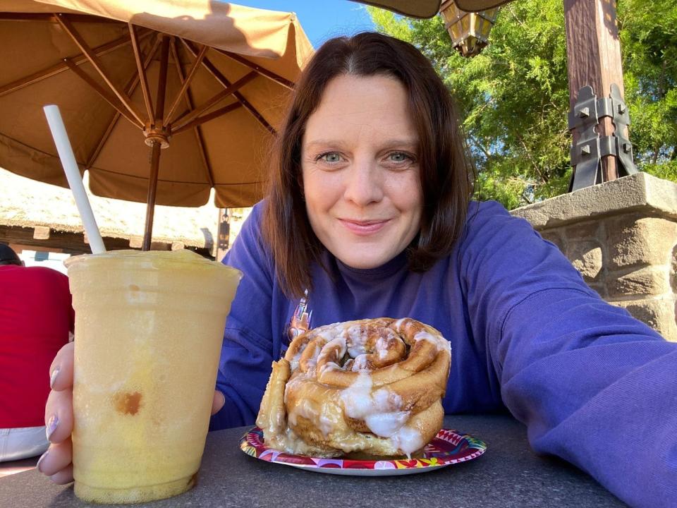 kari posing for a photo with an apple cider slushee and a big cinnamon role from gaston's tavern in disney world