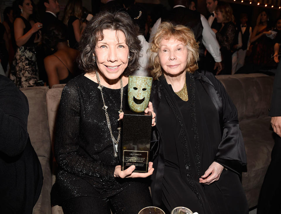 Lily Tomlin and Jane Wagner seated, holding a trophy with a comedy mask. Both are dressed in elegant black attire at an award event