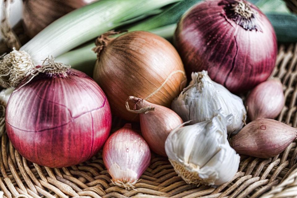 assortment of red and yellow onions displayed in wicker tray alongside shallots, leeks and garlic