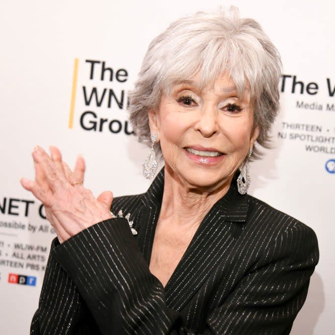 Rita Moreno poses in a pinstripe suit in front of the backdrop of a WNET Group event