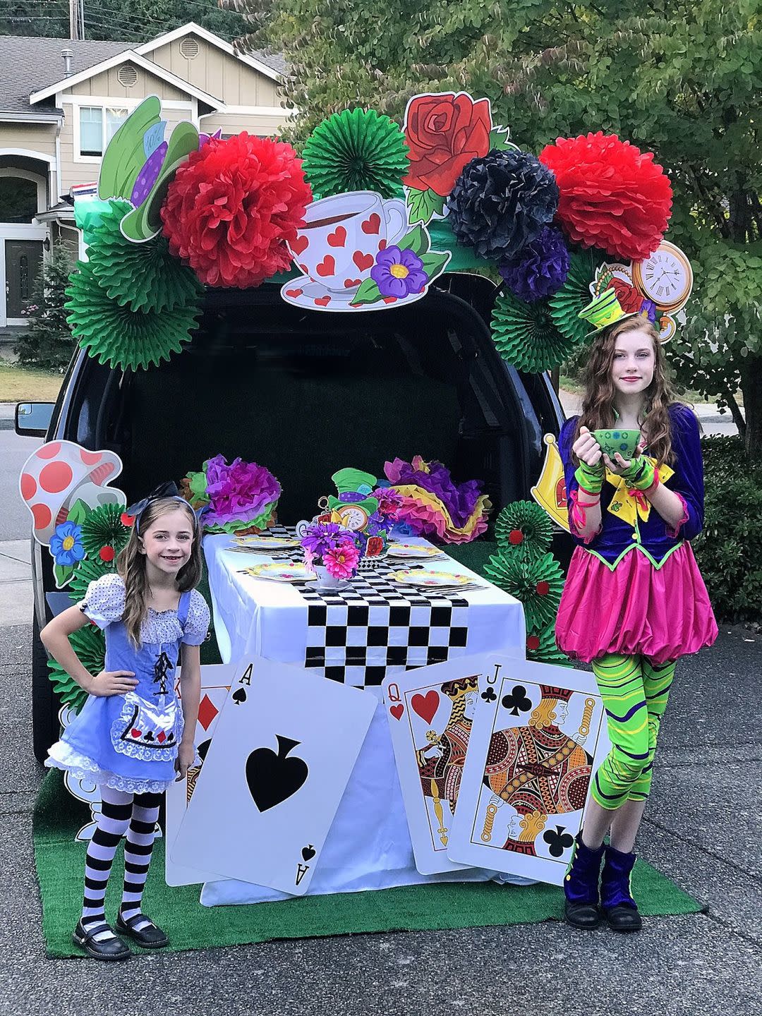 little girl dressesdas alice in wonderland in a blue pinafore dress with black and white striped leggings and older girl dressed as the mad hatter in a purple and pink dress with green leggings standing beside a table in front of an open tailgate with oversized playing cards on the front, a black and white checked tablecloth and over the top of the vehicle are large paper flowers and cut outs of a pocket watch, top hat and tea cup with hearts on it