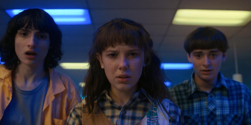 mike, eleven, and will in "Stranger Things 4," looking down at something with shocked expressions on their faces