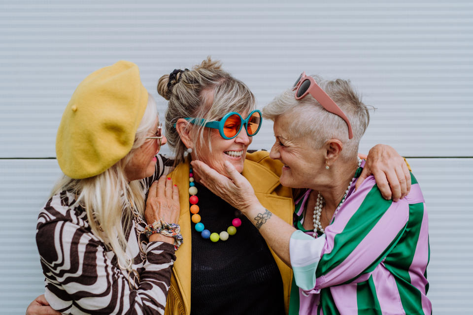 Three women with trendy glasses and colorful clothes smile and hug