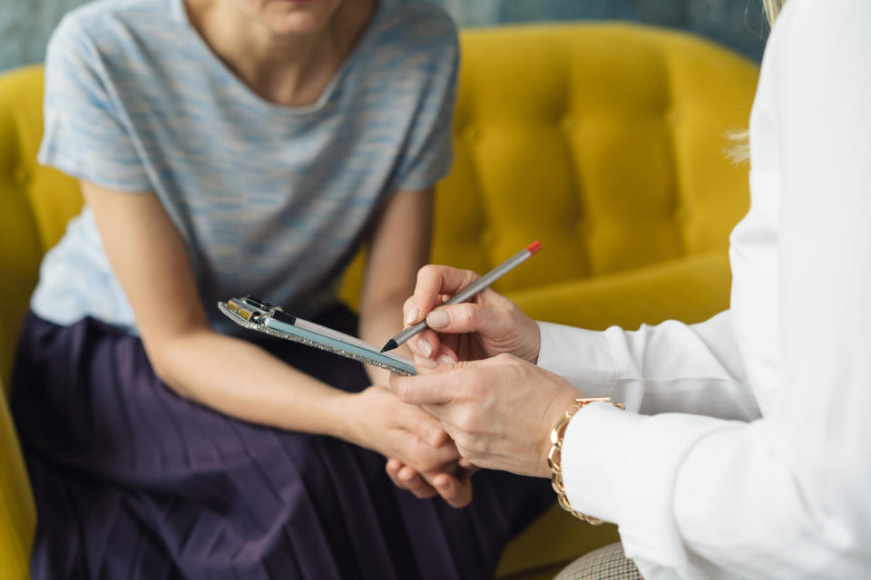 A woman holds a clipboard and pen while talking to another woman who is sitting on a sofa. They appear to be having a serious conversation