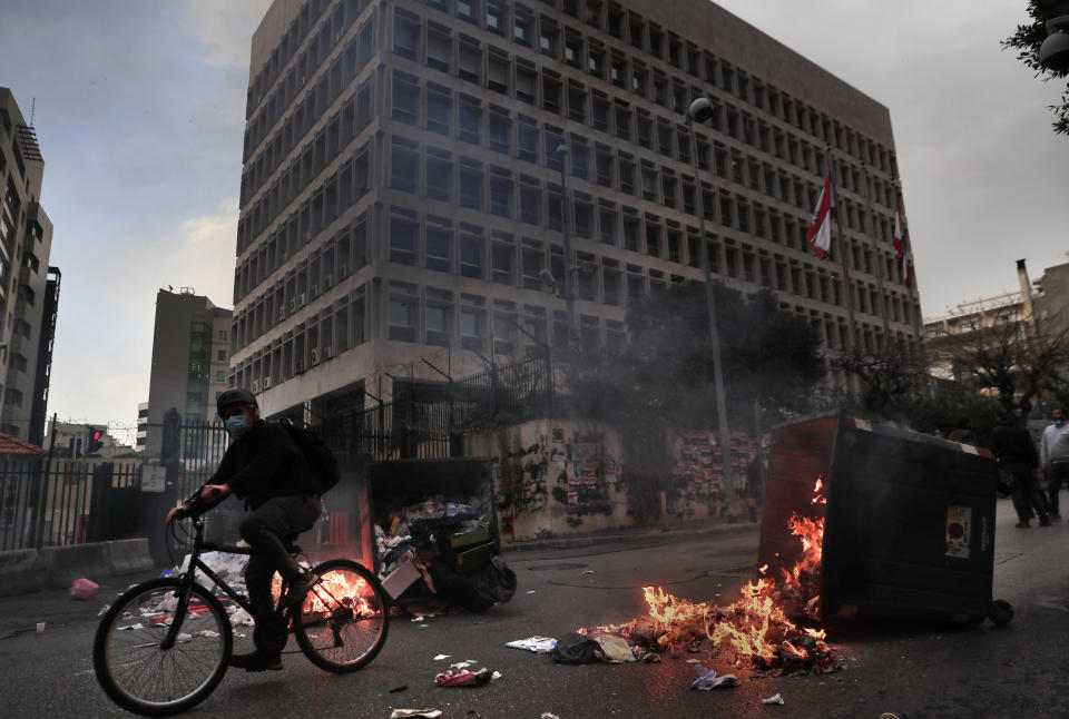 A man rides his bicycle past garbage containers set on fire by protesters blocking a main road in front the Lebanese Central Bank building, background, in Beirut, Lebanon, Monday, March 22, 2021. Protesters blocked some roads in the Lebanese capital with burning tires Monday after talks on the formation of a new Cabinet broke down, heralding more economic and financial collapse for the small Arab country. (AP Photo/Hussein Malla)