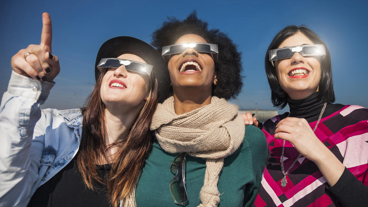  How to watch the solar eclipse: Group of female friends have fun together during a solar eclipse event. Their looking and pointing to the sun wearing the typical glasses normally used to watch a solar eclipse. 