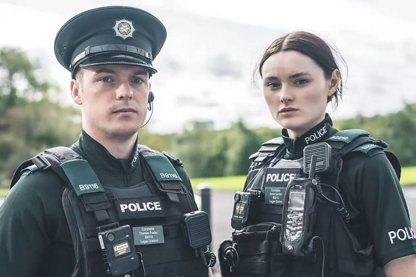 Two young police officers wearing uniform as stood outside as they look into the camera with neutral facial expressions