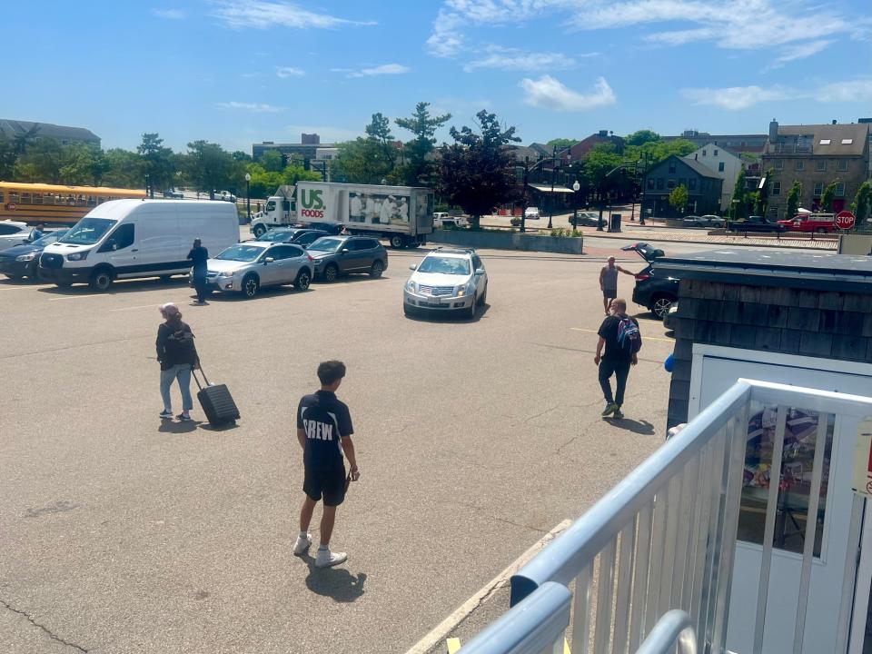 A parking area with several cars, a white van, and a school bus. A man wearing a shirt that says "Crew" stands in the parking lot as a woman with a suitcase and a man with a backpack walk past.