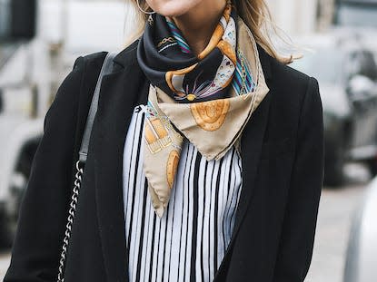 woman on a street wearing yellow sunglasses, a black suit, a stripped shirt, and a colorful silk scarf around her neck