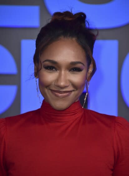 A woman wearing a red turtleneck smiles for cameras at a red carpet event.