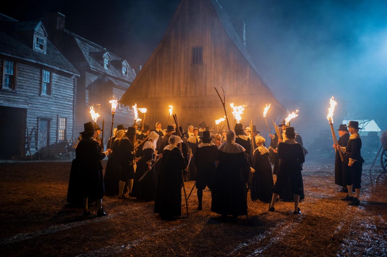 A mob of settlers in traditional black and white pilgrim attire stands in a circle with lighted torches and pitchforks.