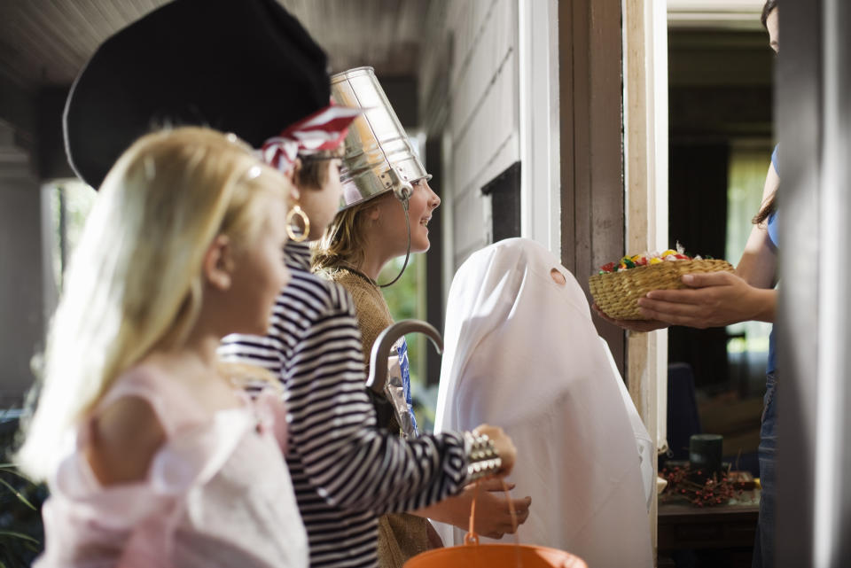 Children dressed in costumes, including a ghost and pirate, trick-or-treating at a house while an adult hands out candy in a basket