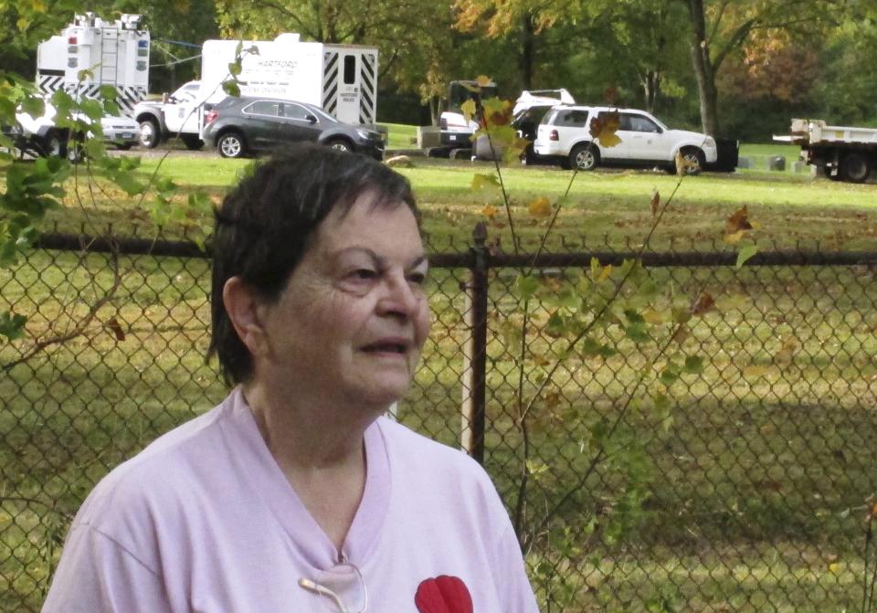 Patricia Congelosi talks with reporters in her backyard as authorities in the abutting Northwood Cemetery, rear, exhume the remains of two unidentified victims of the 1944 Hartford circus fire, Monday, Oct. 7, 2019, in Windsor, Conn. Congelosi, 82, said she was supposed to go to the circus on the day of the fire, but her family went to the beach instead. Officials will try to determine if one of the unidentified victims is Grace Fifield, of Newport, Vt. Grace Fifield was never seen again after attending the circus. (AP Photo/Dave Collins)