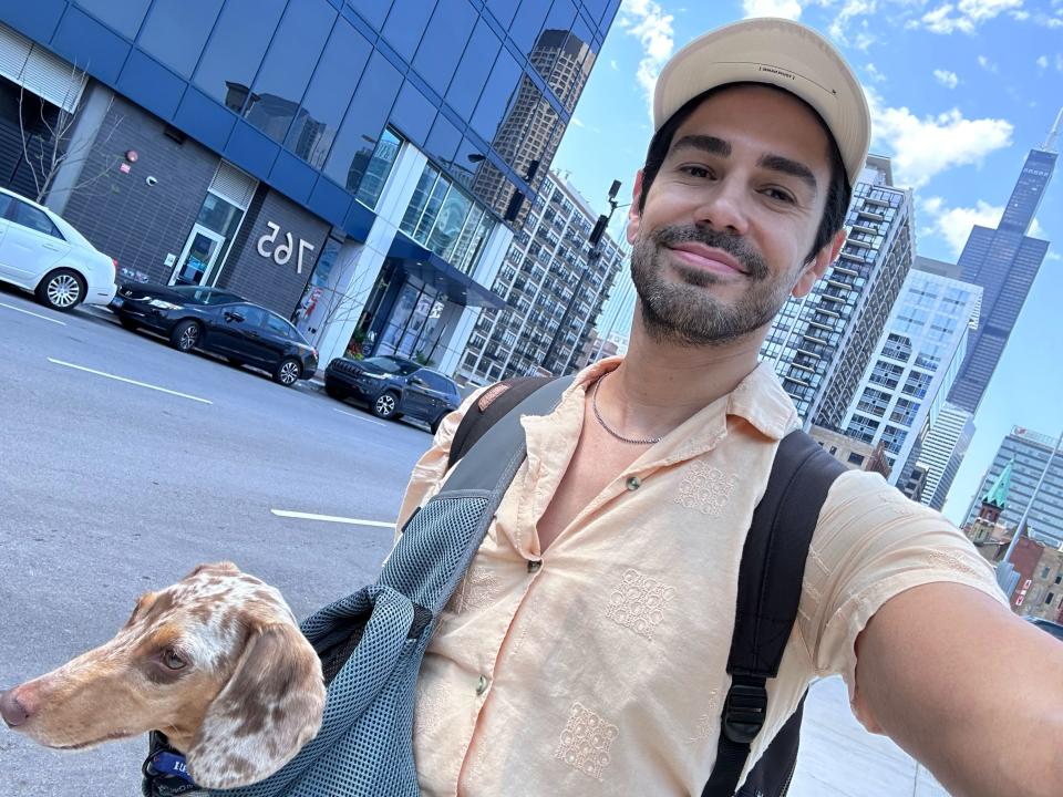 Sean, wearing a cream-colored shirt and a baseball cap, walks through Chicago with his dog Oliver in a carrier.