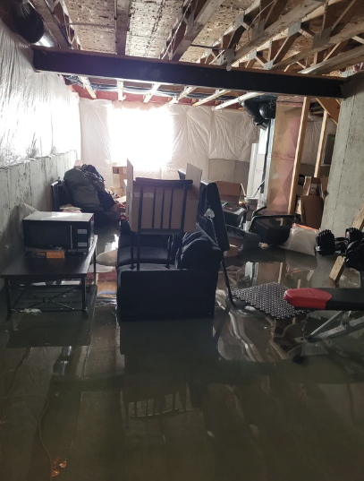 Flooded basement with furniture and items. Water covers the floor. Light shines through an end window, highlighting the extent of flooding