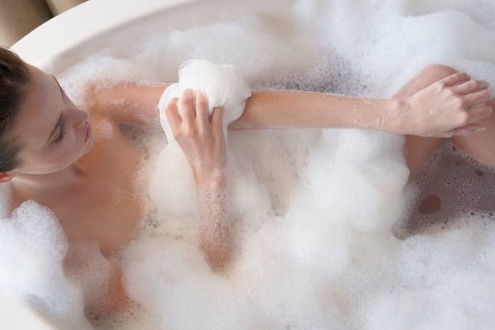 A person relaxes in a bubble bath and scrubs his arm with a loofah