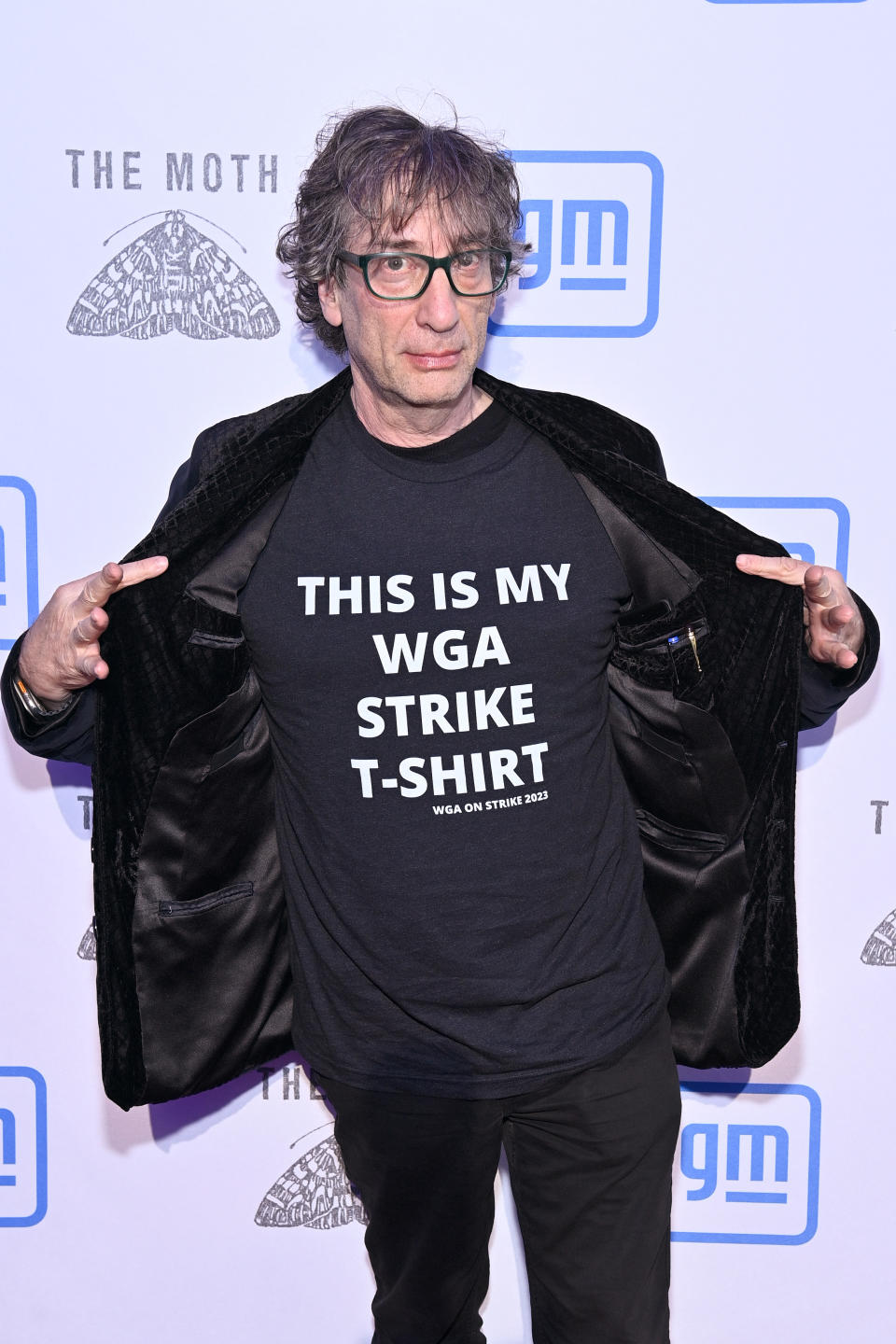 Neil Gaiman, wearing glasses and a black t-shirt that reads "This is my WGA Strike T-Shirt," poses at a media event