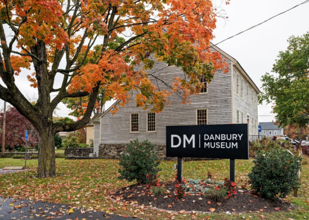 A museum sign in front of a wooden building with a large tree in front of it. 