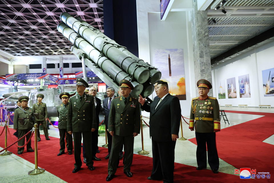 Kim Jong Un and Sergei Shoigu stand on a red carpet among several officials in uniform near posters of rocket launches in a building that appears to house military equipment.