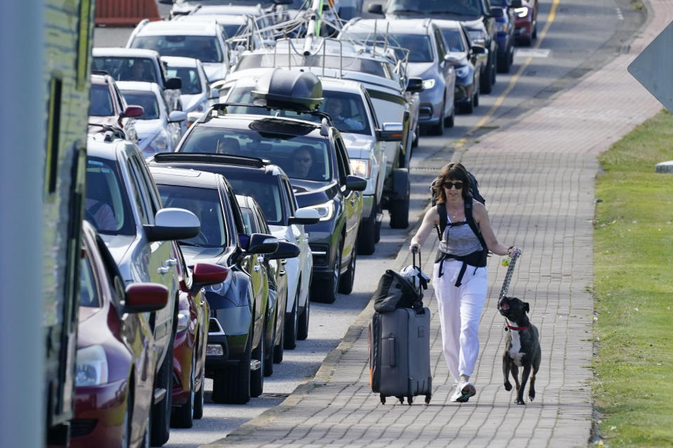 Terri Mills walks with her dog, Carlos, past a line of vehicles waiting to enter Canada at the Peace Arch border crossing Monday, Aug. 9, 2021, in Blaine, Wash. Mills, an American from Grizzly Flats, Calif., was heading to visit her Canadian husband. Canada lifted its prohibition on Americans crossing the border to shop, vacation or visit, but America kept similar restrictions in place, part of a bumpy return to normalcy from coronavirus travel bans. (AP Photo/Elaine Thompson)