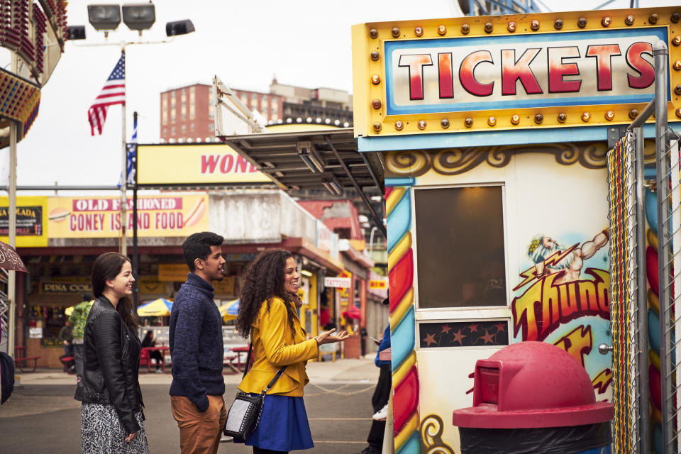 Three people stand in line at a brightly decorated ticket booth at an amusement park. The sign reads "TICKETS"