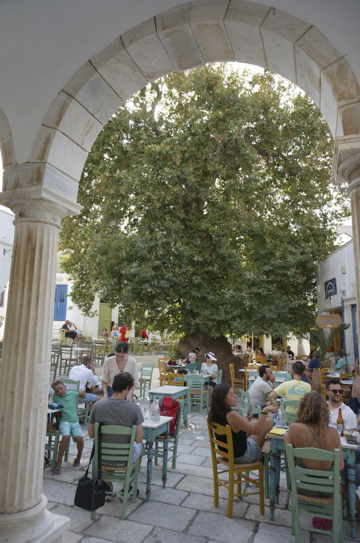A large plane tree shades the café-lined, marble-paved central square in the village of Pyrgos on Tinos island, Greece, on Aug. 26, 2021. Tinian villages are richly decorated in marble from local quarries, and Pyrgos houses the marble craft museum as well as many artist workshops. (AP Photo/Giovanna Dell’Orto)