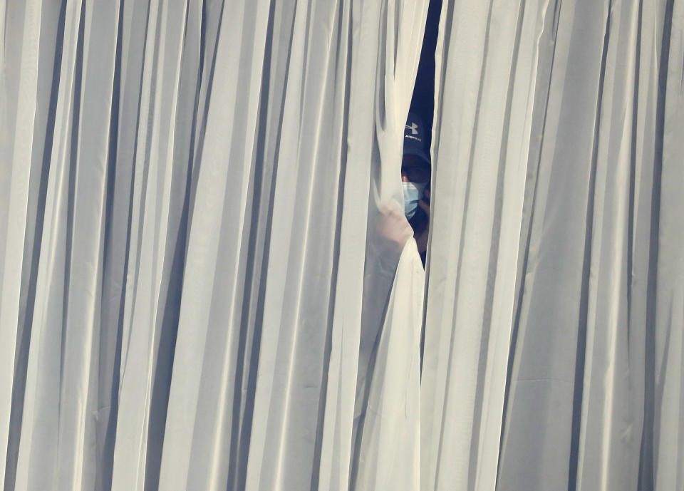 <p>A man wearing a mask against the coronavirus peers out from behind the net curtain in his room at the Radisson Blu Edwardian Hotel, near Heathrow Airport, London, Monday Feb. 15, 2021, where people will remain during a 10 day quarantine period after returning to England from one of 33 "red list" countries. New regulations now in force require anyone who has been in a 'high-risk' location to enter England through a designated port and have pre-booked a package to stay at one of the Government's managed quarantine facilities. (AP Photo/Alastair Grant)</p>
