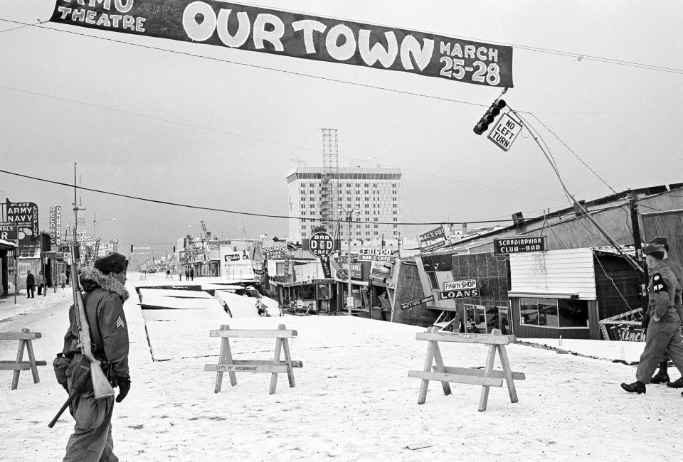 File - In this March 29, 1964 file photo, a soldier crosses Fourth Avenue, the main street in Anchorage, Alaska, a city under martial law following the devastation visited upon it by an earthquake. The sign overhead advertises a production of the Thornton Wilder play. Buildings at right were called a total loss. North America's largest earthquake rattled Alaska 50 years ago, killing 15 people and creating a tsunami that killed 124 more from Alaska to California. The magnitude 9.2 quake hit at 5:30 p.m. on Good Friday, turning soil beneath parts of Anchorage into jelly and collapsing buildings that were not engineered to withstand the force of colliding continental plates. (AP Photo/File)