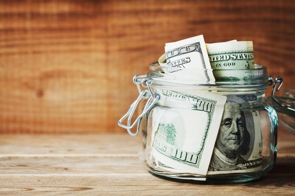 A jar full of $100 bills sitting on a wooden desk.