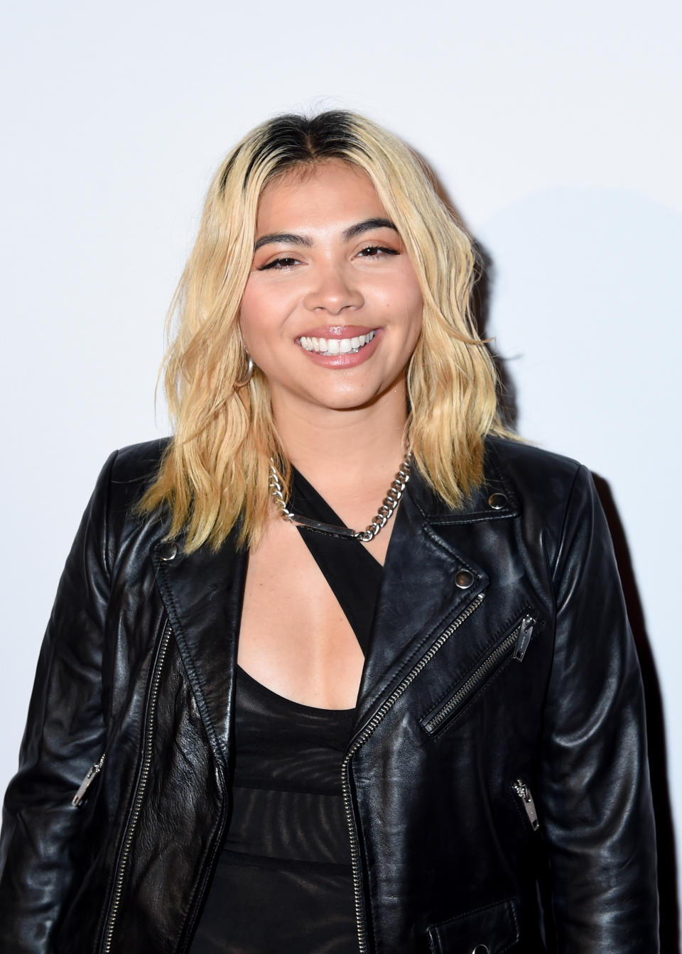 Hayley Kiyoko smiles, wearing a black leather jacket over a black top, with a chain necklace