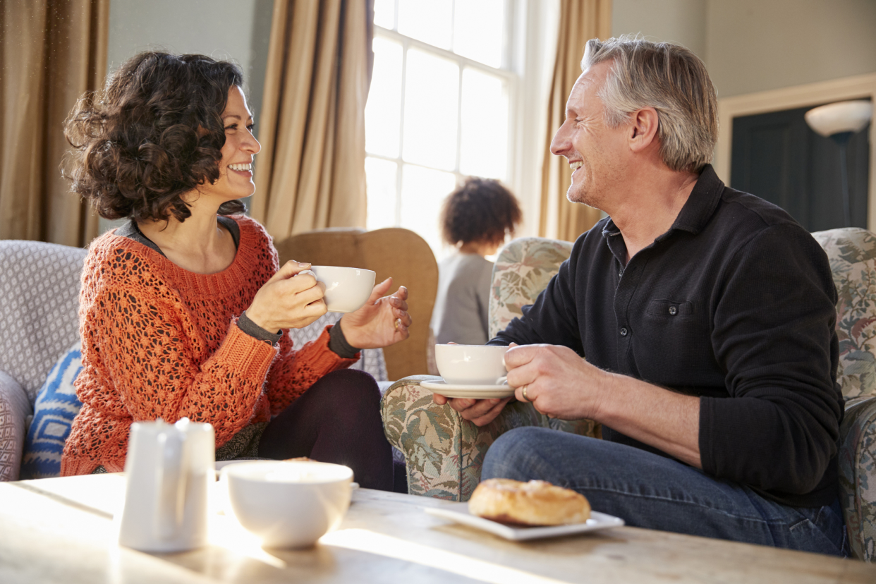 Middle-aged couple sitting down in upholstered chairs, having coffee, in a coffee house, on a date, near a table with two plates of danishes, with window, wall, and a person at a table in the background