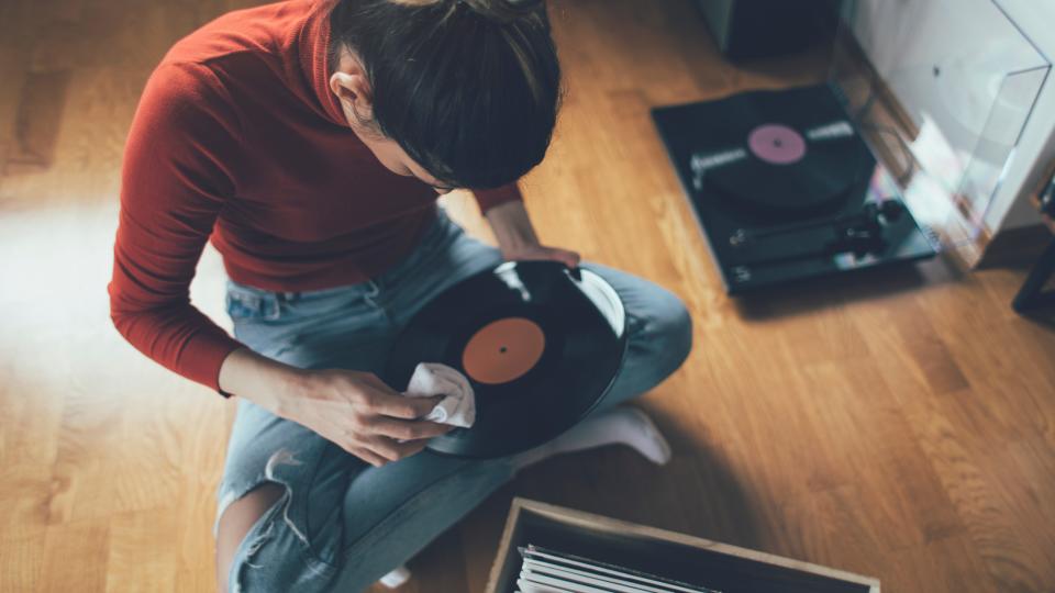 A woman sitting cross-legged, cleaning a vinyl record with a cloth, in a bedroom