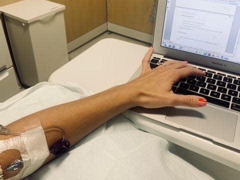 Photo of Emily Holi's left arm typing on a laptop, while in a hospital bed. She has an IV in the crease of her arm and her nails are painted red. The laptop rests on a small table and behind this, you can see a plastic garbage can and beige curtain. 