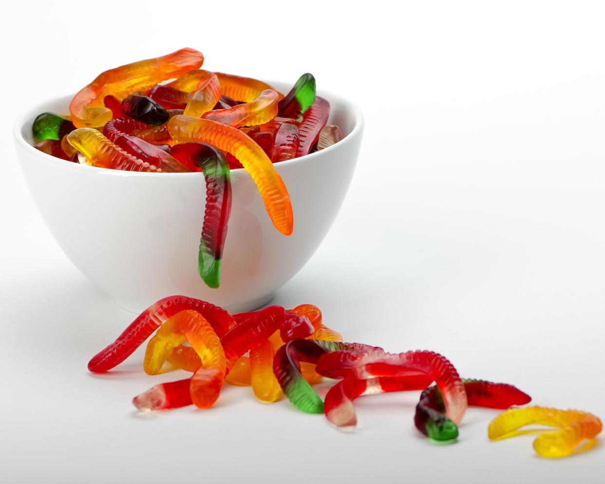 A white bowl of sugar-free gummy worms with a trail of gummy worms spilling onto a white table with a white background