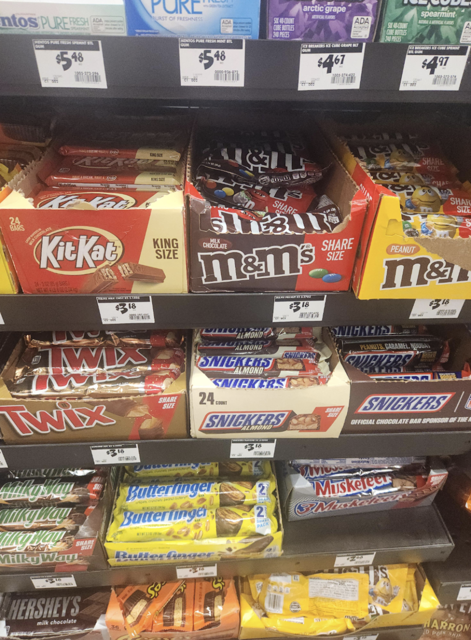 Store shelf filled with various candy bars including KitKat, M&M's, Twix, Snickers, Butterfinger, 3 Musketeers, Hershey's, and others