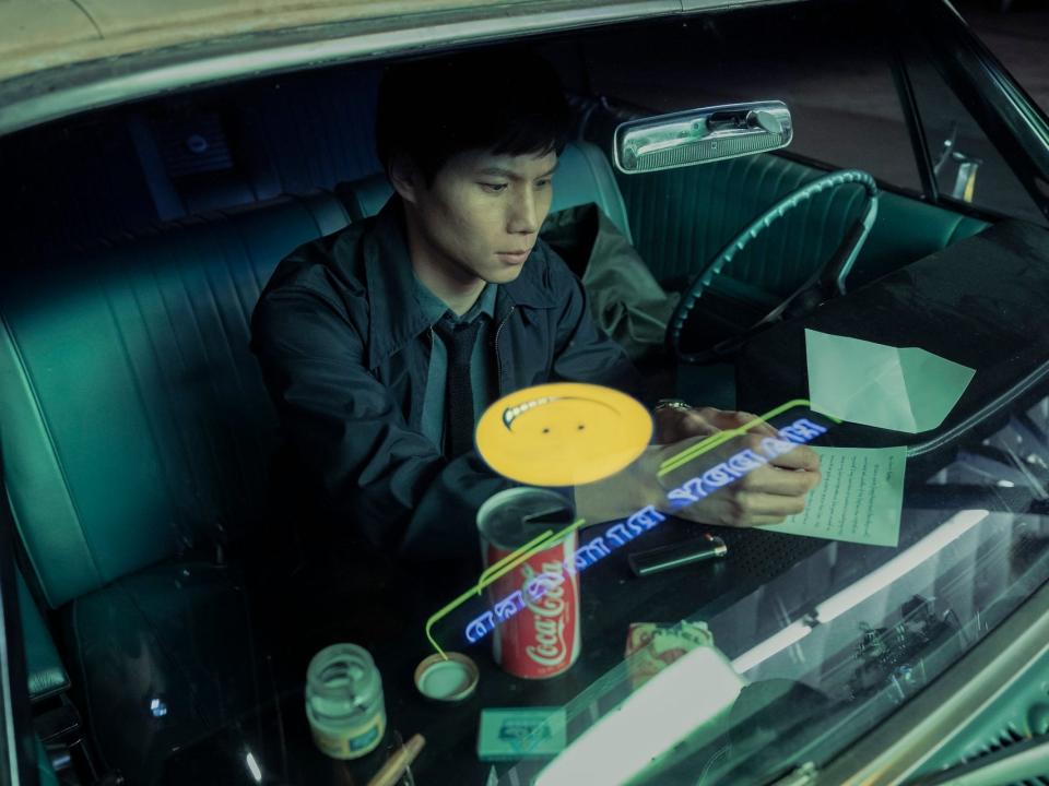 hoa xuande in the sympathizer, writing on a piece of paper on his dashboard while sitting in a car. there's a smiley face reflected on his windshield