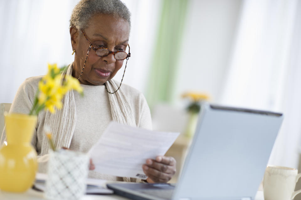 An older woman wearing glasses is looking at a document while sitting at a table with a laptop and flower vase. She appears focused on her work