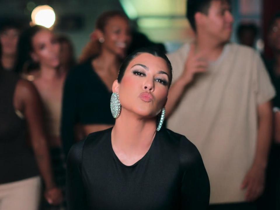 Kourtney Kardashian purses her lips and poses in front of a crowd of roller skaters. she's wearing large, circular, sparkling earrings