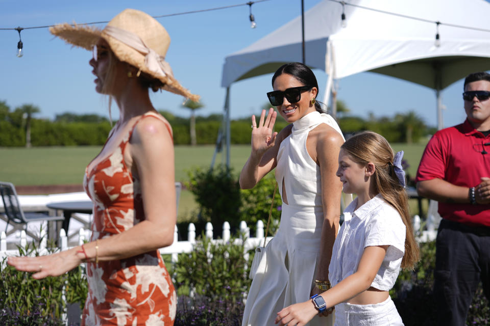 Meghan Markle, Duchess of Sussex, waves to the press as she walks with Delfina Blaquier, left, and Alba Figueras, the wife and daughter of Argentine professional polo player Ignacio "Nacho" Figueras, as she arrives for the 2024 Royal Salute Polo Challenge to Benefit Sentebale, Friday, April 12, 2024, in Wellington, Fla. Prince Harry, co-founding patron of the Sentebale charity, will play on the Royal Salute Sentebale Team. (AP Photo/Rebecca Blackwell)